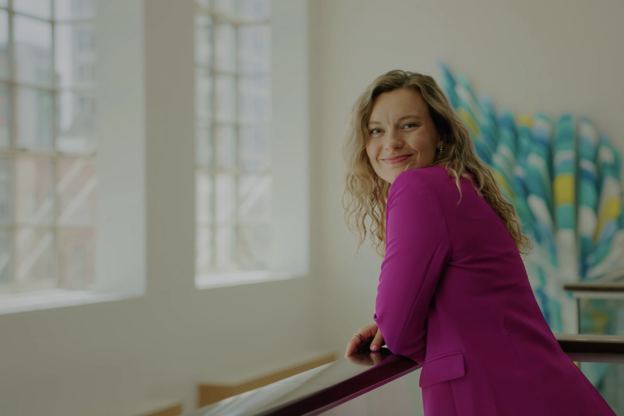 Rachel Eslick smiling and leaning on a railing in a brightly lit space, wearing a magenta blazer.